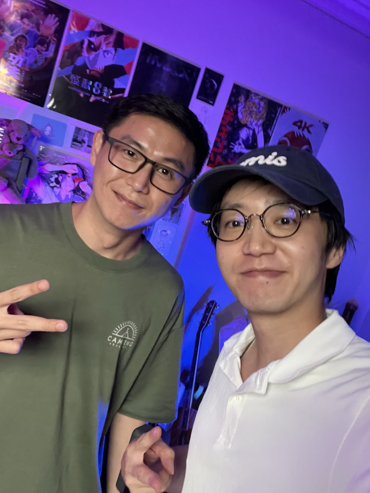 藍紫燈光的工作室裡兩個人並肩合照,其中一人比出手勢,背景是牆上的海報。